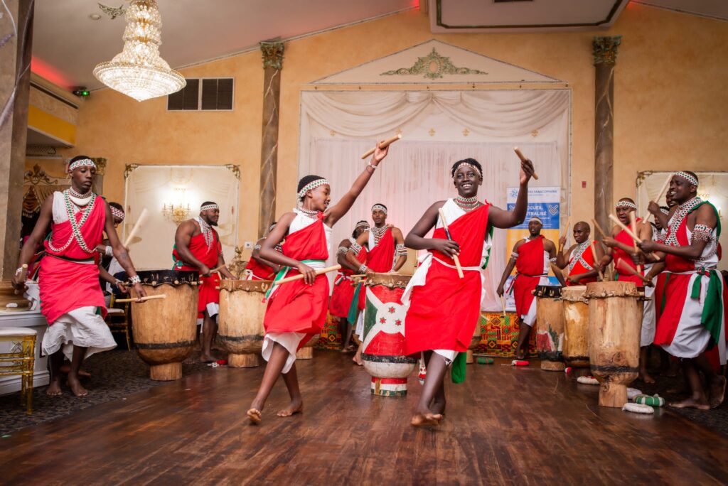 The Remesha Drums ensemble mid-performance, drummers in red and white regalia around carved wooden drums